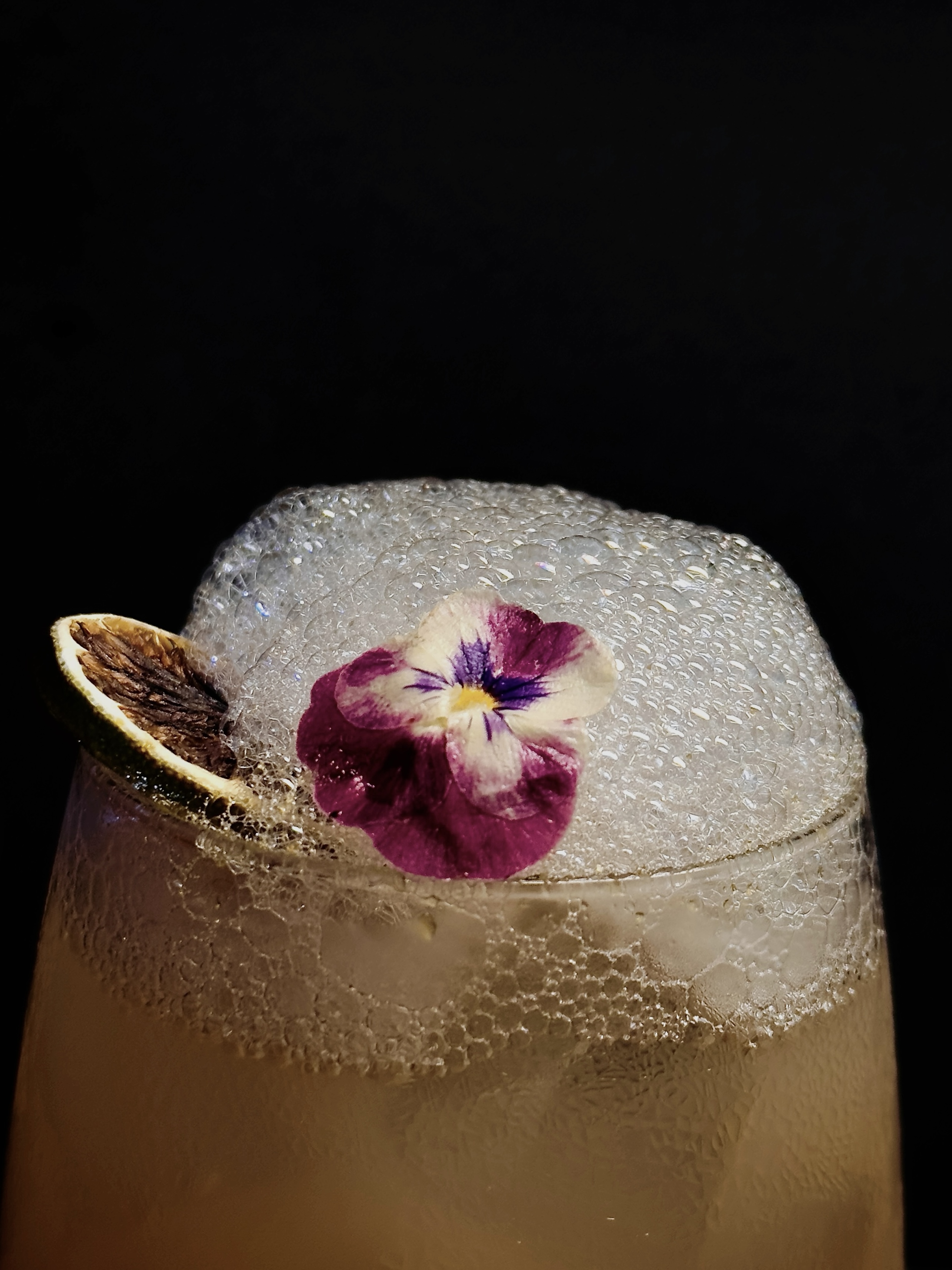 Cocktail with frothy white head, edible pansy and dehydrated lime garnish on a dark background at Dissolved Solids