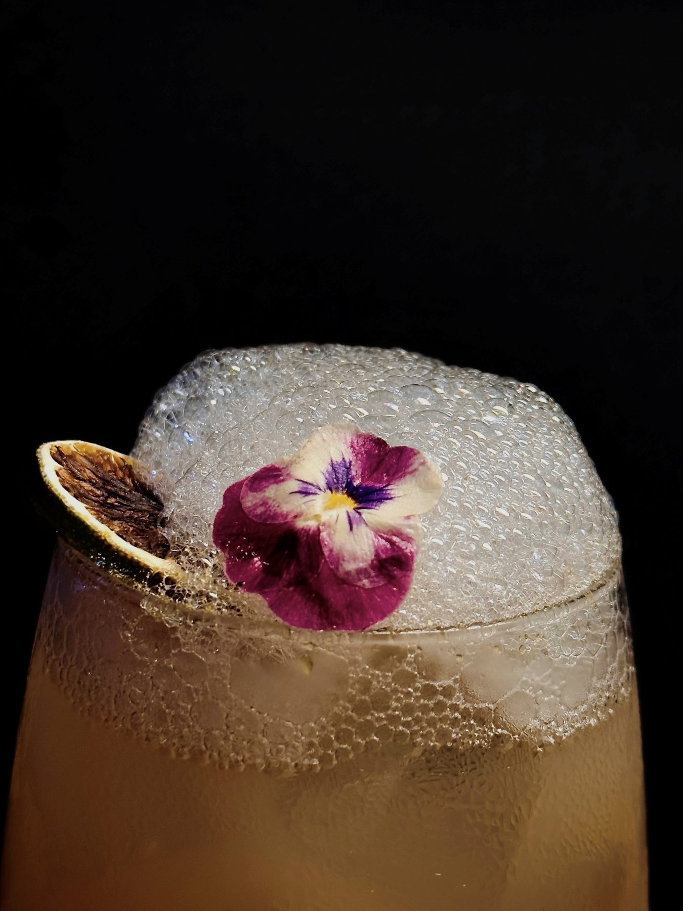 Cocktail with a frothy white head, edible pansy and dehydrated lime garnish on a dark background at Dissolved Solids
