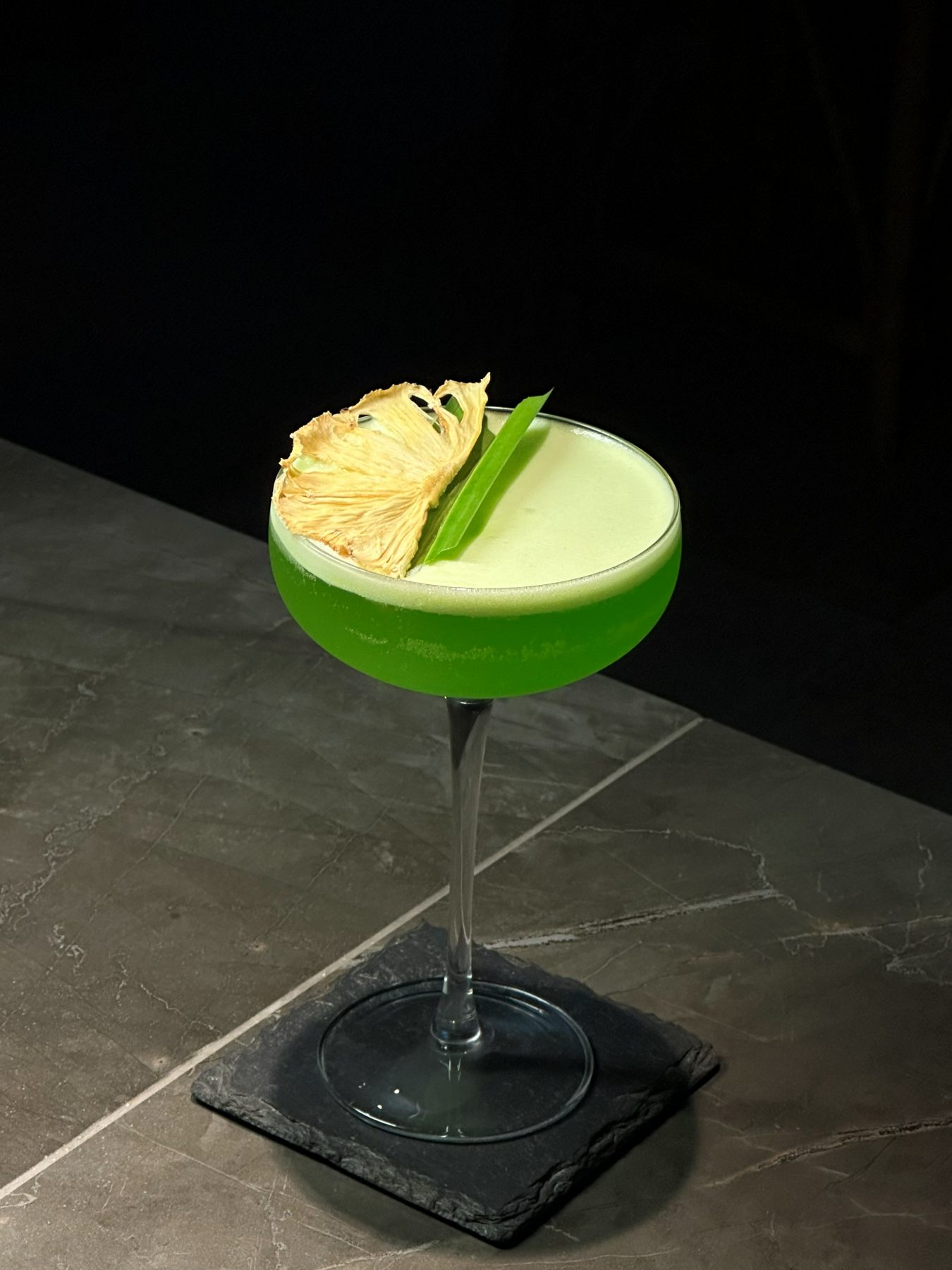 Bright green coupe cocktail with dehydrated pineapple wedge and pandan-leaf garnish on dark slate at Dissolved Solids Damansara Kim