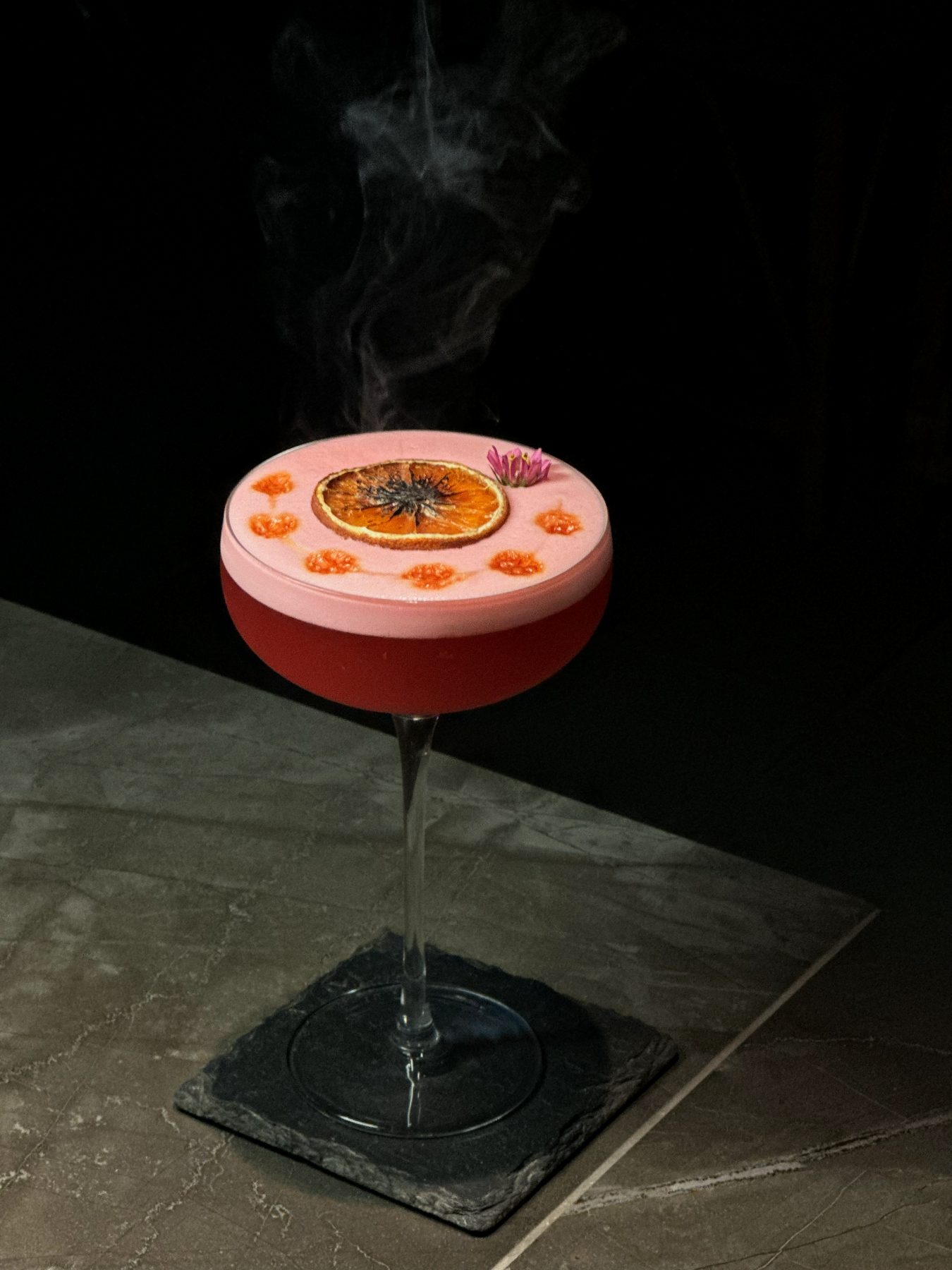 Pink coupe cocktail with dried orange wheel, edible flower and rising smoke at Dissolved Solids