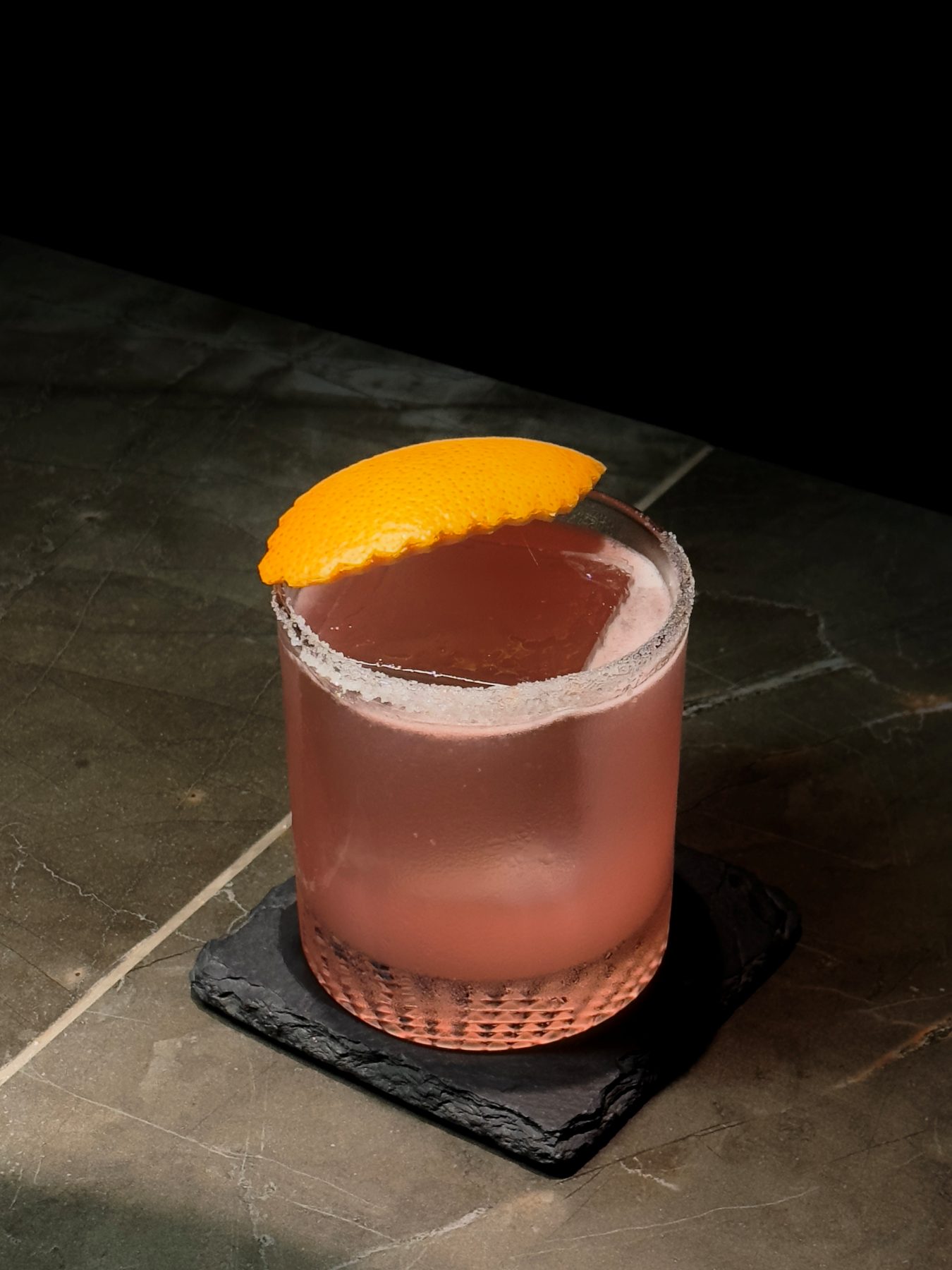 Pink-orange rocks cocktail with a salt rim and an expressed orange peel on slate at Dissolved Solids
