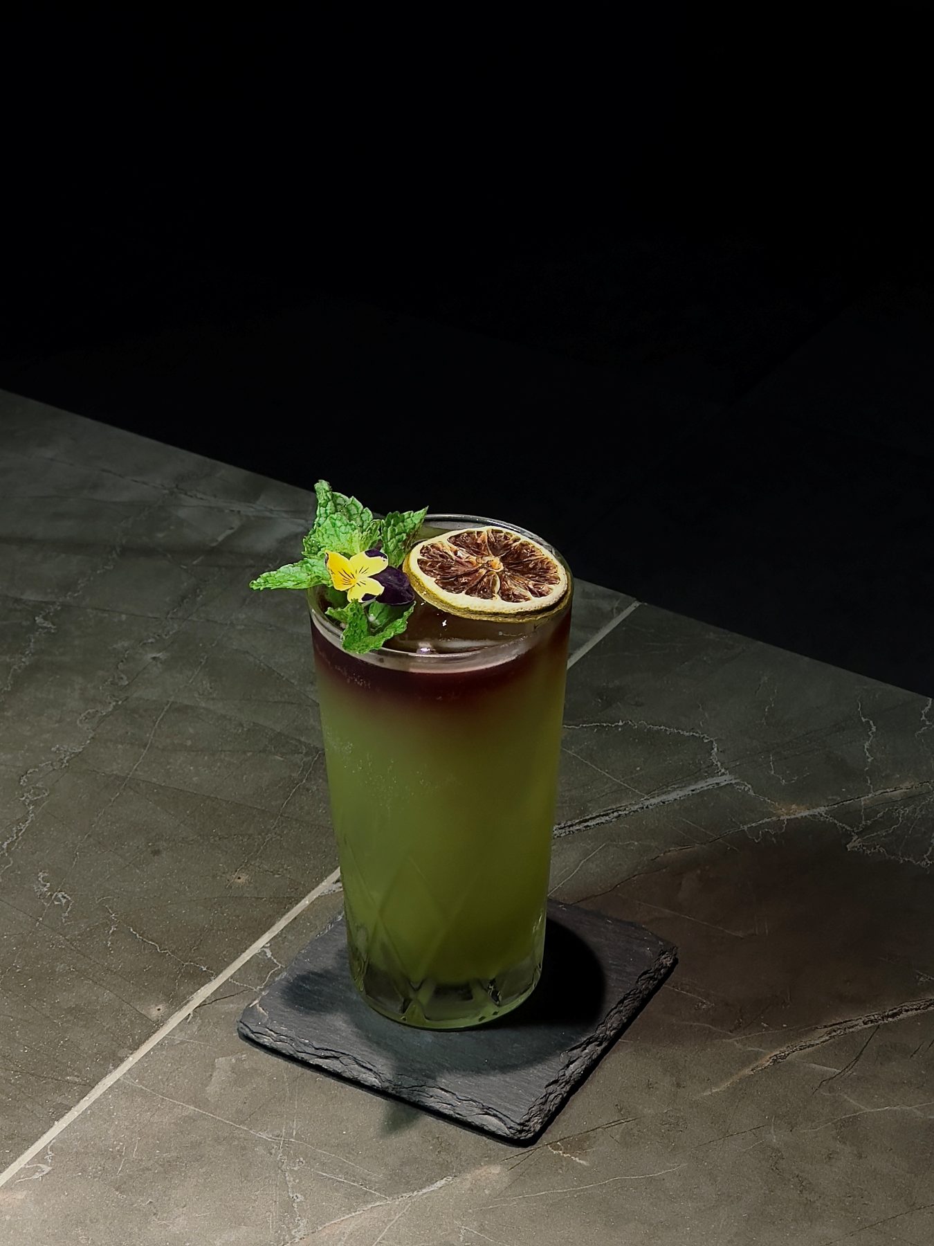 Tall green highball cocktail with mint, edible pansy and dehydrated lime garnish on slate at Dissolved Solids