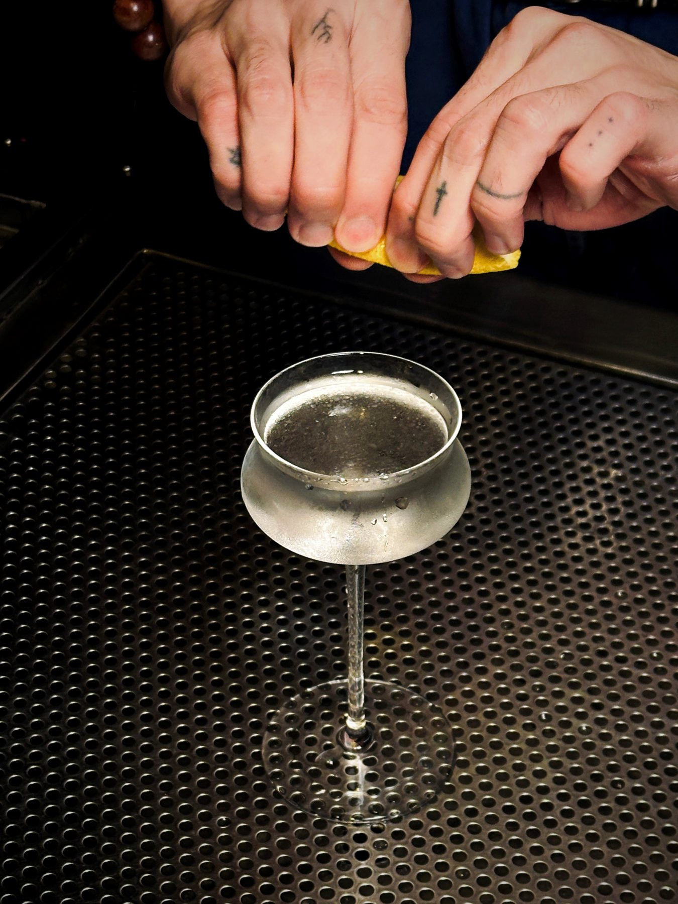 Tattooed hands expressing a lemon peel over a chilled coupe at Soluble Solids