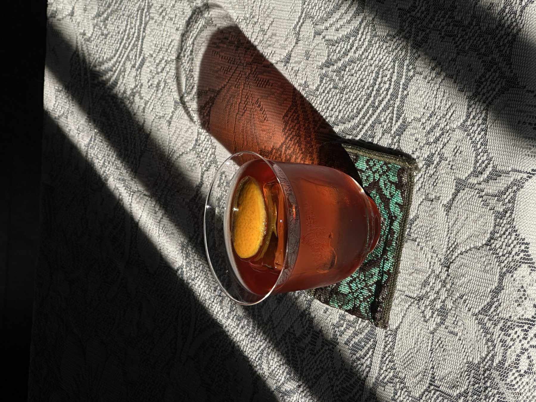Amber rocks cocktail with a carved wood garnish on patterned lace, harsh midday shadow at Soluble Solids